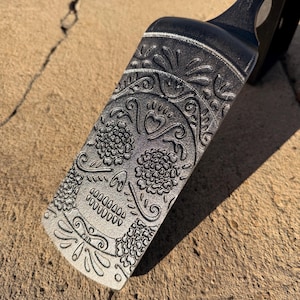 May include: A black and silver cake server with a detailed sugar skull design. The sugar skull is intricately carved with floral and heart motifs. The handle is black, and the server is resting on a concrete surface.