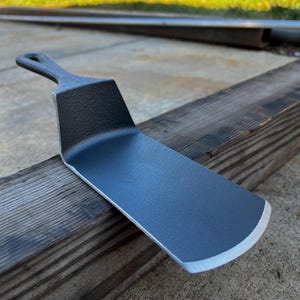 May include: A dark gray metal spatula with a rounded edge and a handle with a hole. The spatula is resting on a weathered wooden surface. The spatula is designed for cooking.