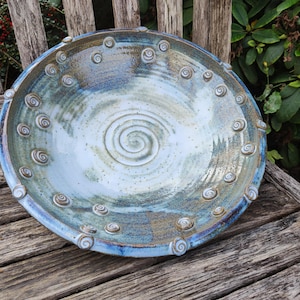 Könnte beinhalten: Eine runde Keramikschale mit einer blau-weißen Glasur und einem markanten Spiralmuster in der Mitte. Der Rand ist mit zahlreichen kleinen, erhabenen Tonspiralen verziert, die kreisförmig um den Rand der Schale angeordnet sind.