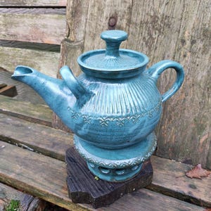 Könnte beinhalten: Türkisgrüne Keramik-Teekanne mit Deckel und Henkel, auf einem dunklen Holzsockel. Die Teekanne hat vertikale Rippendetails und ein dekoratives Blumenmuster. Der Ausguss ist gebogen und der Griff gewölbt. Die Teekanne fasst etwa 1,2 Liter.