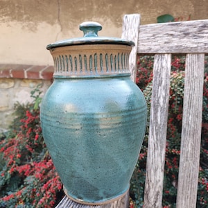 Op de afbeelding: Een handgemaakte keramische pot met deksel, voorzien van een glanzend blauwgroen glazuur over een gespikkelde klei. Het bovenste gedeelte van de hals vertoont een reeks verticale, uitgesneden groeven die de natuurlijke beige klei onthullen.
