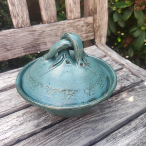 Op de afbeelding: Een teal-kleurige keramische kom met deksel, een glanzende afwerking en een gespikkelde textuur. Het deksel heeft een gedraaide lusvormige handgreep in het midden en een delicaat reliëf bloemmotief dat de koepel omcirkelt.