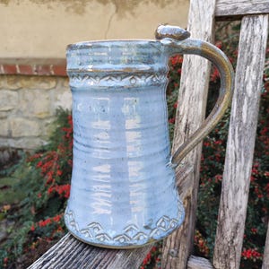 Könnte beinhalten: Ein hoher, zylindrischer Keramikbecher mit hellblauer Glasur und glänzender Oberfläche. Der Becher verfügt über einen geschwungenen Henkel und dekorative geprägte geometrische Muster an den oberen und unteren Rändern sowie eine Textur.