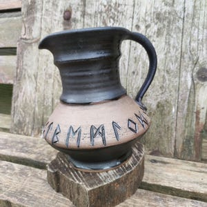 Könnte beinhalten: Ein schwarzer Keramik-Krug mit einem braunen Band und Runen um die Mitte. Der Krug steht auf einem Holzsockel.