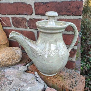 Può includere: Una teiera in ceramica verde con un coperchio e un manico. La teiera ha una glassa macchiata e un disegno di foglie intorno al corpo.