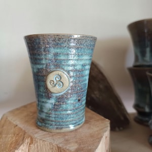 Op de afbeelding: Een handgemaakte keramische beker met een turquoise en bruin glazuur. De beker heeft een Keltisch triskelesymbool en wordt tentoongesteld op een houten blok. Het ontwerp heeft een rustiek, ambachtelijk gevoel.
