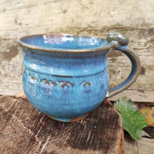Könnte beinhalten: Eine handgefertigte Keramiktasse mit türkisfarbener Glasur. Die Tasse hat einen abgerundeten Körper, einen gebogenen Henkel und ein dekoratives Band mit geprägten Details. Der Rand und der Henkel sind dunkler braun. Die Tasse ist etwa 10 cm hoch.