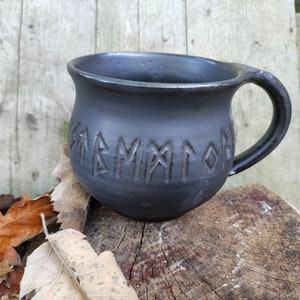 Könnte beinhalten: Schwarze Keramiktasse mit abgerundeter Form und Henkel. Die Tasse hat Runen, die in die Seite geschnitzt sind.