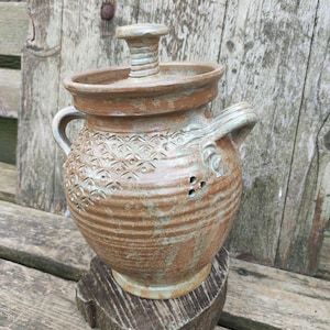 Pode incluir: Um jarro de cerâmica castanho artesanal, com tampa e asa. O jarro apresenta uma superfície texturizada com um padrão decorativo e pequenos orifícios. É exibido sobre uma base de madeira contra um fundo de madeira envelhecida.