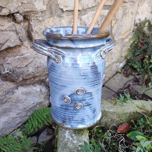 Może przedstawiać: Cylindryczny ceramiczny pojemnik na przybory kuchenne z jasnoniebieską i białą glazurą, wyposażony w dwa boczne uchwyty oraz małe spiralne akcenty dekoracyjne. Wewnątrz pojemnika znajdują się trzy drewniane przybory kuchenne.