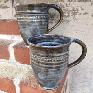 Può includere: Due tazze in ceramica fatte a mano dall'aspetto rustico e strutturato. Le tazze sono principalmente marroni con accenti blu e bianchi, con una fascia decorativa al centro. Ogni tazza ha un grande manico curvo.
