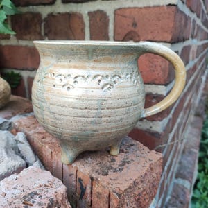 Puede incluir: Una taza de cerámica hecha a mano con un esmalte beige y azul claro. La taza tiene un cuerpo redondeado, un asa y tres pequeños pies. El borde está decorado con un patrón floral. La taza está sobre un ladrillo rojo.