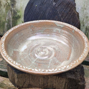 Może przedstawiać: Ręcznie robiona ceramiczna miska w jasnobrązowych i beżowych odcieniach. Miska ma dekoracyjne obramowanie i spiralny wzór na środku. Miska stoi na drewnianej powierzchni.