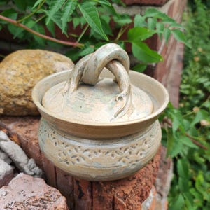 May include: A round ceramic lidded jar with a beige and sage green glaze. The lid features a sculptural, knotted handle. The body of the jar is decorated with a repeating embossed floral pattern around the centre, and the rim is slightly flared.