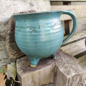 Op de afbeelding: Een turquoise keramische mok met een handvat en drie kleine voetjes. De mok heeft een brede opening en een decoratief golfpatroon rond de bovenkant. De mok staat op een houten oppervlak.