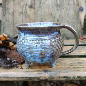 Könnte beinhalten: Eine handgefertigte Keramiktasse mit einem einzigartigen Design. Die Tasse hat eine blau-braune Glasur, einen abgerundeten Körper, einen Henkel und drei kleine Füße. Die Außenseite hat ein strukturiertes, gemustertes Design. Geeignet für heiße oder kalte Getränke.