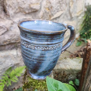 Könnte beinhalten: Ein Keramikbecher mit blau-brauner Glasur, der im oberen Bereich ein horizontales Band mit geprägten kreisförmigen Mustern aufweist. Der Becher hat einen ausgestellten Rand, einen geschwungenen Henkel und einen Fuß, was ihm ein texturiertes Aussehen verleiht.