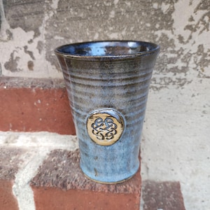Könnte beinhalten: Ein handgefertigter Keramikbecher mit blauer und brauner Glasur. Der zylindrische Becher hat eine strukturierte Oberfläche und ein dekoratives Emblem. Der Becher ist etwa 13 cm hoch und 8 cm im Durchmesser.