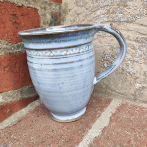 Puede incluir: Una taza de cerámica hecha a mano con un esmalte azul claro. La taza presenta un cuerpo redondeado, un asa resistente y una banda decorativa cerca de la parte superior. El borde y el asa tienen un acento azul más oscuro. La taza mide aproximadamente 10 cm de alto.