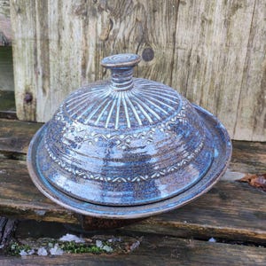 Peut inclure: Un beurrier en céramique bleu avec un couvercle. Le plat a une base ronde et un couvercle en forme de dôme avec une petite poignée. Le couvercle et la base sont décorés d'un motif de lignes et de points. Le plat est posé sur une surface en bois.