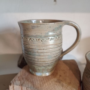 Puede incluir: Una taza de cerámica hecha a mano con un esmalte marrón claro y verde azulado. La taza presenta líneas horizontales y una banda decorativa de motivos florales. Tiene un asa curva y se apoya sobre una superficie de madera.
