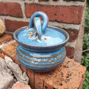 Puede incluir: Un tarro de cerámica hecho a mano con tapa y asa, esmaltado en tonos azules y marrones. El tarro presenta un patrón texturizado alrededor de la base y un asa en forma de lazo en la tapa. El tarro está sobre una superficie de ladrillo.