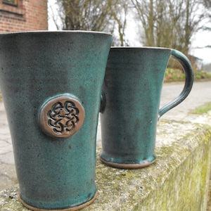 Könnte beinhalten: Zwei türkisfarbene Keramikbecher mit rustikalem Finish. Ein Becher hat einen Henkel, der andere ein dekoratives Emblem. Die Becher werden im Freien auf einer Steinmauer präsentiert, was auf ein handwerkliches Thema hindeutet.