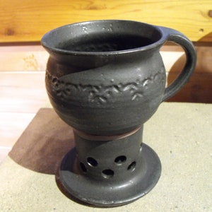 Könnte beinhalten: Eine dunkelgraue Keramiktasse mit Henkel, die auf einem passenden Sockel mit kreisförmigen Aussparungen steht. Die Tasse hat ein dekoratives Blumenmuster um ihren Mittelteil. Das Design deutet auf eine rustikale, handwerkliche Ästhetik hin, die für Getränke geeignet ist.