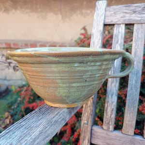 Peut inclure: Un bol mélangeur en céramique avec un bord large et une poignée latérale unique. Le bol présente un vernis moucheté dans des tons de vert sauge et de brun terreux, avec des stries horizontales visibles sur la surface et une base arrondie.