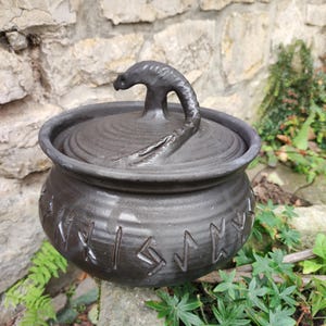 Può includere: Un barattolo in ceramica grigio scuro con coperchio, corpo arrotondato e bordo largo. Il coperchio presenta un manico scolpito a forma di testa di creatura curva. L'esterno del barattolo è decorato con una fascia di simboli runici incisi.