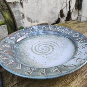 Peut inclure: Assiette en céramique faite à la main, glaçure bleu clair et marron. Le bord présente des runes sculptées et le centre un motif en spirale. L'assiette mesure environ 25 cm de diamètre, idéale pour servir des aliments ou comme pièce décorative.