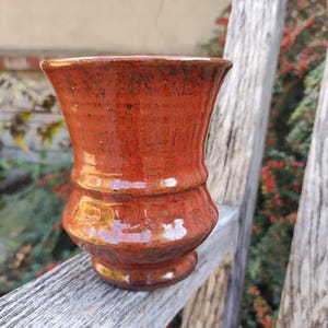 Könnte beinhalten: Ein Keramikbecher mit einer glänzenden, leuchtend orangefarbenen Glasur, die feine dunkle Sprenkel und eine strukturierte Oberfläche aufweist. Der Becher hat einen weit ausgestellten Rand, eine verjüngte Mitte und einen abgerundeten Boden.