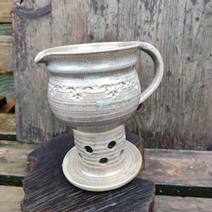 Könnte beinhalten: Eine handgefertigte Keramik-Sahnekännchen mit Ausguss und Henkel. Der cremefarbene Krug hat ein dekoratives Blumenmuster in der Mitte und steht auf einem Sockel mit kreisförmigen Aussparungen. Die Keramik ist mit Blau- und Brauntönen glasiert.