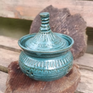 Puede incluir: Un tarro de cerámica turquesa con tapa, superficie texturizada y patrones decorativos. El tarro tiene una base redonda con pequeños agujeros y una tapa cónica con un asa texturizada. El tarro se exhibe sobre una superficie de madera.