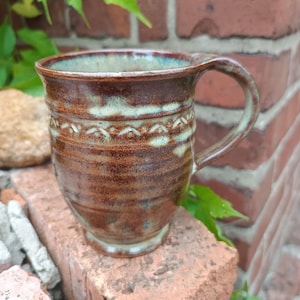 Peut inclure: Une tasse en céramique avec un vernis brillant brun et sarcelle, présentant une bande horizontale texturée avec un motif d'arc répétitif sur la partie supérieure, un bord évasé et une anse incurvée, avec une finition artisanale.