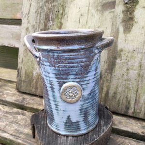 Könnte beinhalten: Eine blaue und braune Keramikvase mit einem keltischen Knotendesign in Gold. Die Vase hat zwei Henkel und eine strukturierte Oberfläche.