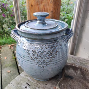 Op de afbeelding: Een lichtblauwe keramische pot met bijpassend deksel en twee handvatten aan de zijkanten. De pot heeft een gespikkelde glazuurafwerking en een driehoekig textuurpatroon aan de voorzijde. Het deksel heeft een centrale knop en een donkerblauwe rand.
