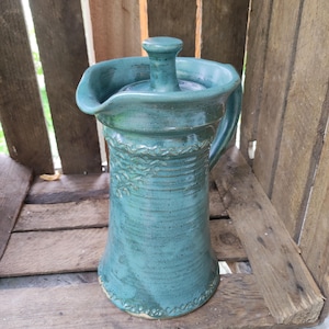 May include: A teal ceramic pitcher with a matching lid, featuring a flared rim, a side handle, and a textured band of leaf-like patterns around the top and bottom sections of the cylindrical body, finished with a glossy, mottled glaze.