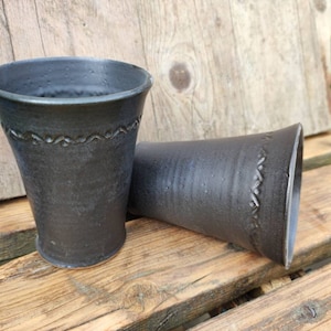 Könnte beinhalten: Zwei schwarze Keramikbecher mit strukturierter Oberfläche und einem dekorativen, wellenartigen Muster am Rand. Die Becher werden auf einer rustikalen Holzoberfläche präsentiert, was ihre handwerkliche Qualität hervorhebt.