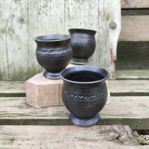 Puede incluir: Tres tazas de cerámica negra con un diseño texturizado. Las tazas están sobre una superficie de madera.