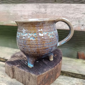 Op de afbeelding: Een bruine keramische mok met een blauw en wit bloemenmotief. De mok heeft drie poten en een handvat. Hij staat op een houten oppervlak.