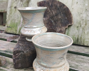 May include: Two handmade ceramic vases with a light beige and blue color scheme. The vases have a flared top, a textured body, and a decorative floral pattern near the base. They are displayed on a weathered wooden surface.