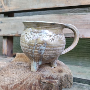 Könnte beinhalten: Eine rustikale, braune und weiße Keramiktasse mit Henkel und drei Beinen. Die Tasse hat ein dekoratives Design mit kleinen Blumen um den oberen Rand.