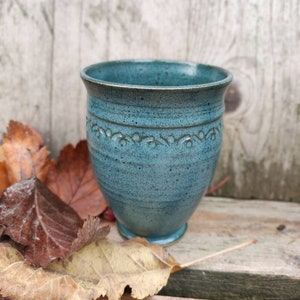 May include: A teal blue ceramic mug with a decorative design around the middle. The mug is sitting on a wooden surface with autumn leaves.