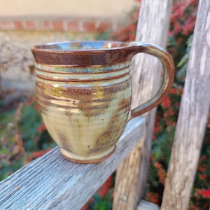 Puede incluir: Una taza de cerámica con cuerpo redondeado y asa curva. El exterior presenta un esmalte reactivo con una mezcla de tonos tierra marrones, beige y sutiles azul verdoso, caracterizado por estrías horizontales y un acabado liso y brillante.