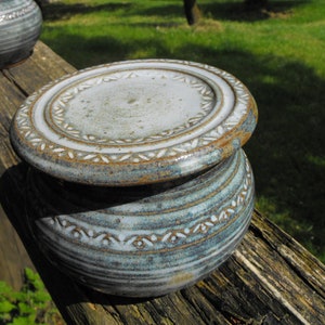 May include: A handmade ceramic container with a lid. The container is a light blue-green color with a white and brown floral pattern around the base and lid. The container is sitting on a weathered wooden surface.
