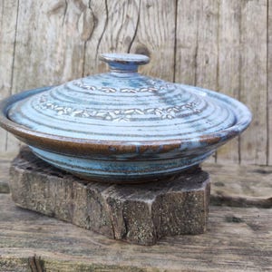 Może przedstawiać: Ręcznie robiona, niebieska ceramiczna miska z pokrywką i kwiatowym wzorem. Brzeg miski jest brązowy i stoi na drewnianej podstawie. Tło to zwietrzała drewniana powierzchnia.