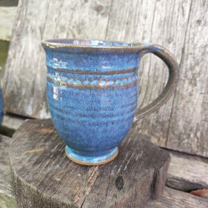 May include: A handmade blue ceramic mug with a brown handle and base. The mug has a textured surface with horizontal lines and two brown bands. The mug is sitting on a wooden surface.