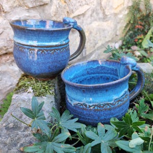 Könnte beinhalten: Zwei Keramikbecher mit einer leuchtend blauen Glasur und dunkelblauen sowie braunen Akzenten. Jeder Becher hat einen abgerundeten Körper, einen geschwungenen Henkel mit einer kleinen Daumenauflage oben und ein geprägtes Muster in der Mitte.