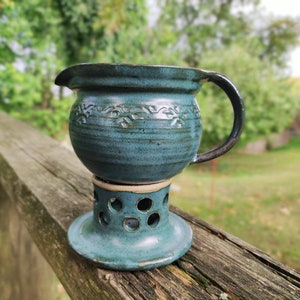 Może przedstawiać: Ręcznie robiony turkusowy ceramiczny dzbanek z ciemnym uchwytem i kwiatowymi detalami. Dzbanek stoi na pasującej turkusowej podstawie z okrągłymi wycięciami. Element jest wystawiony na zewnątrz na drewnianej powierzchni.